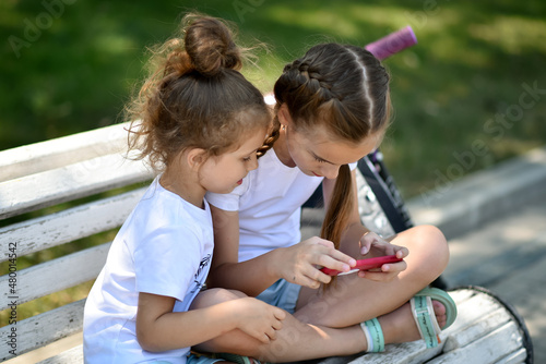 Two sister girls sit on a bench and communicate on a gadget.