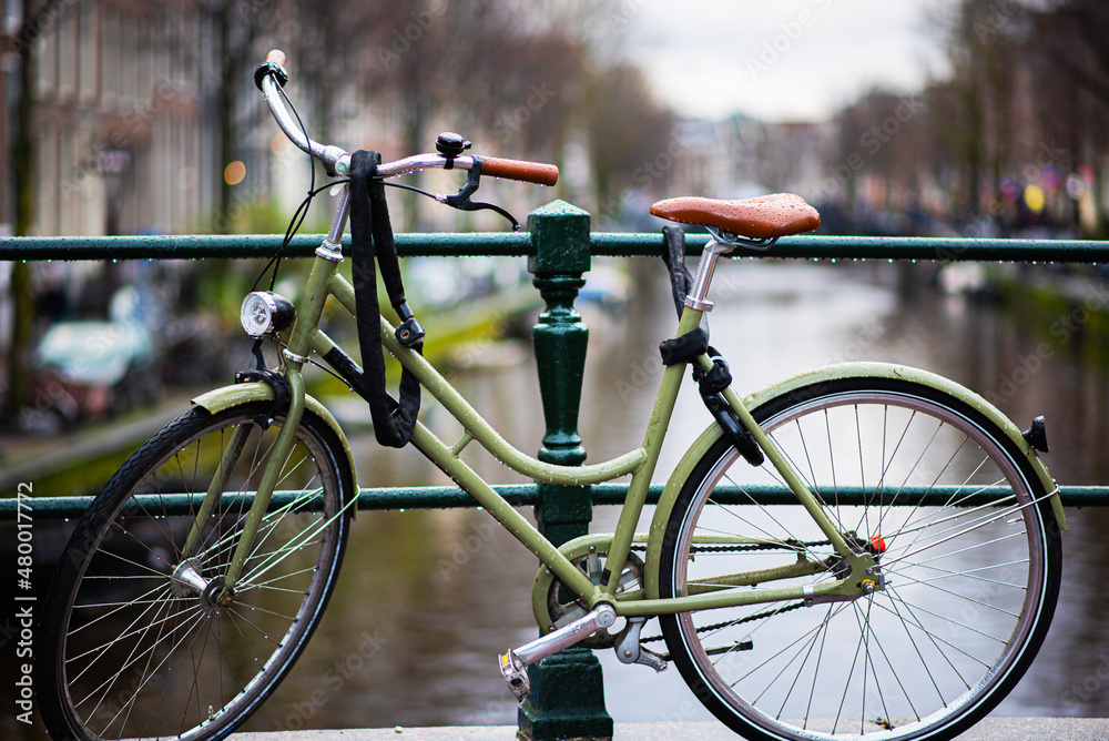 Fototapeta premium Rainy day in Amsterdam. Bicycle on wet street in Amsterdam in rain. Bike over canal. Picturesque town landscape in Netherlands. Toned photo.
