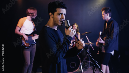 young singer performing song with blurred music band on stage.