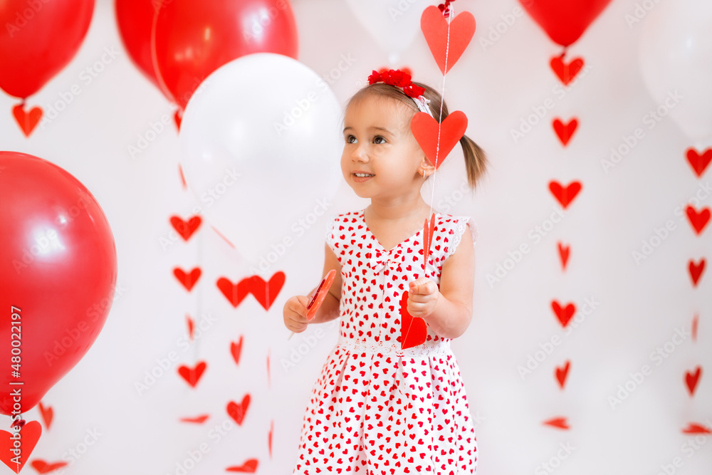 The child holds in his hand a garland with red hearts and a balloon. Bright decorations for Valentine's Day. Dress with a pattern of many hearts