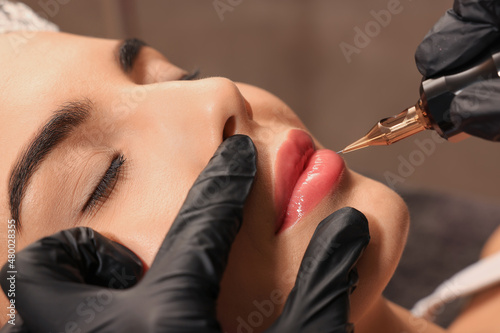 Tela Young woman undergoing procedure of permanent lip makeup in tattoo salon, closeu