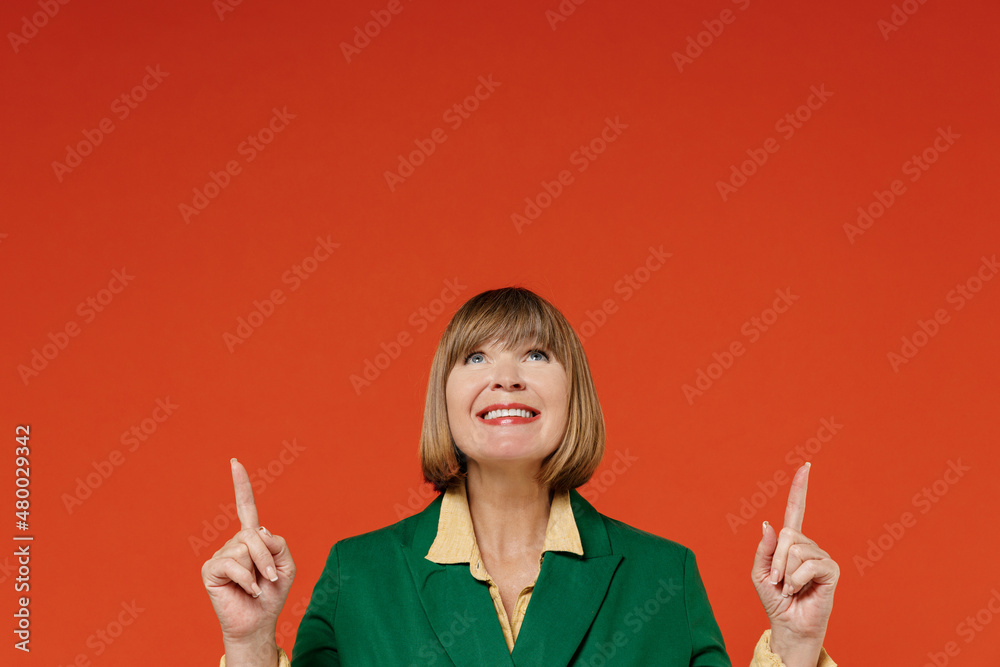 Elderly woman 50s wear green classic suit point index finger overhead on workspace area mock up copy space isolated on plain orange color background studio portrait. People business lifestyle concept. Elderly woman 50s wear green classic suit point index finger overhead on workspace area mock up copy space isolated on plain orange color background studio portrait. People business lifestyle concept.