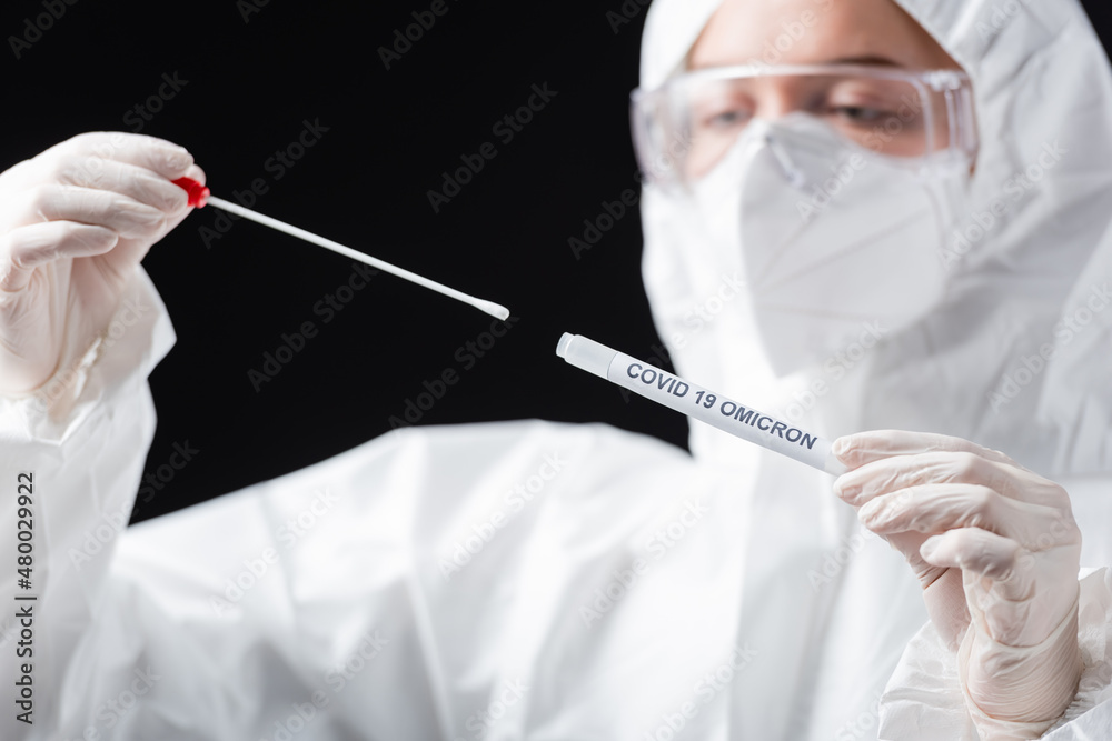 blurred scientist in hazmat suit holding covid-19 omicron variant swab test isolated on black. blurred scientist in hazmat suit holding covid-19 omicron variant swab test isolated on black.