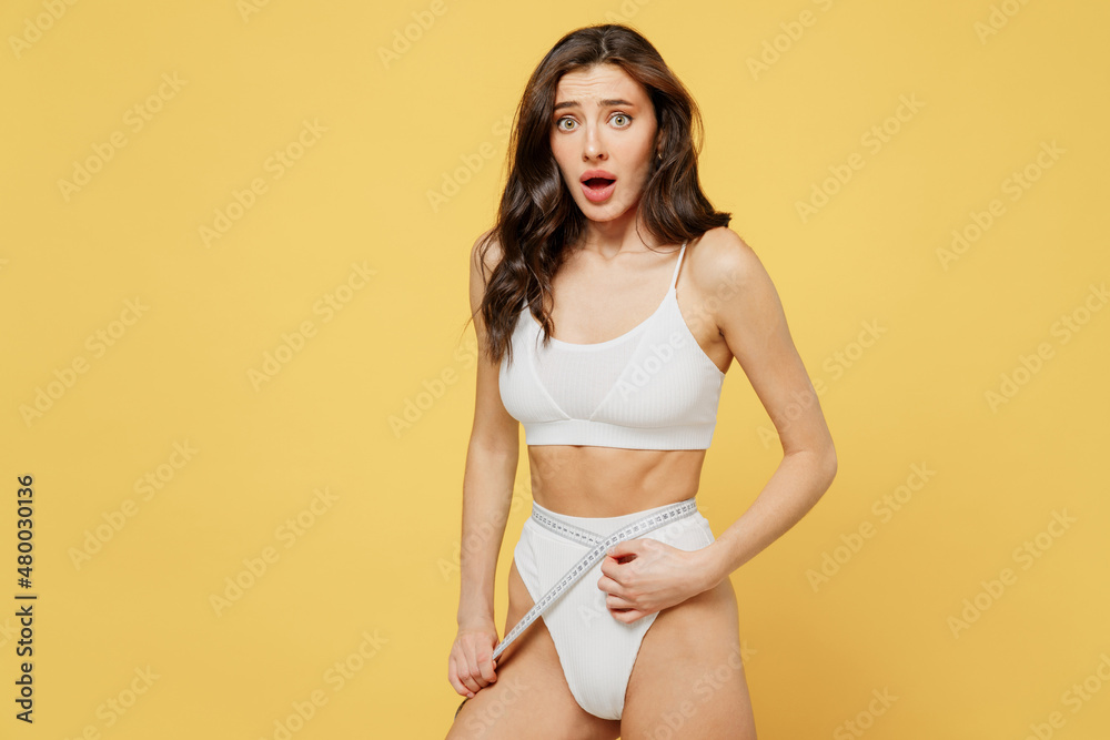 Shocked fun confused displeased young brunette woman 20s wear white underwear with perfect fit body standing posing hold measure tape on waist look camera isolated on plain yellow color background