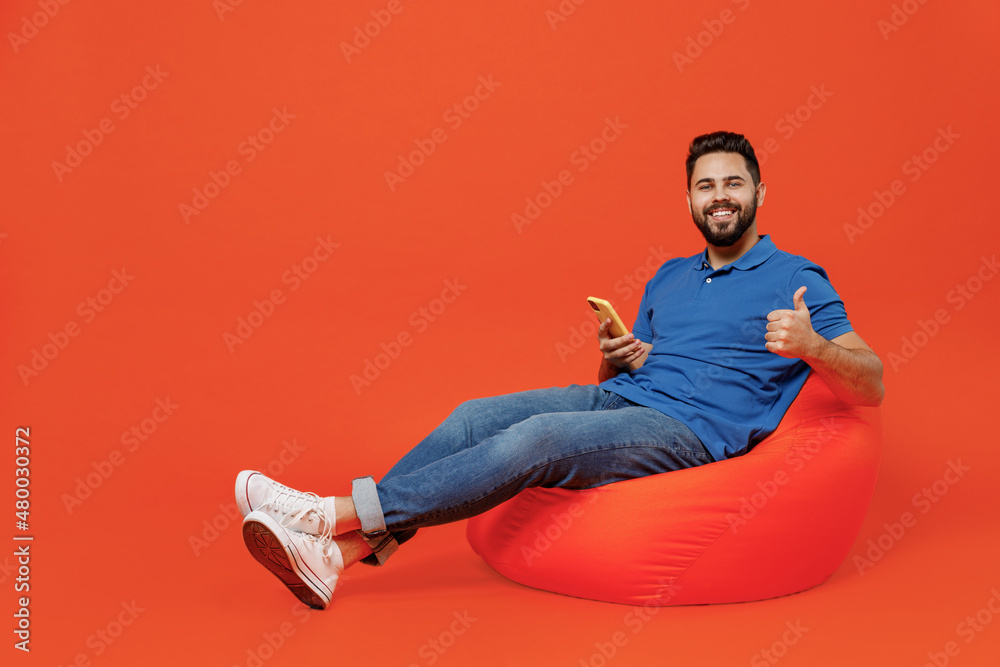 Full body young smiling happy man wear blue t-shirt sit in bag chair hold in hand using mobile cell phone show thumb up isolated on plain orange background studio portrait. People lifestyle concept.