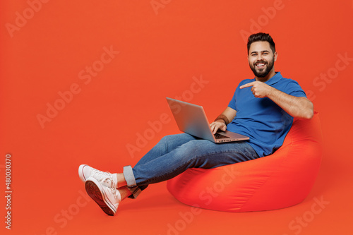 Full body young smiling happy fun man wear basic blue t-shirt sit in bag chair hold use work point on laptop pc computer isolated on plain orange background studio portrait. People lifestyle concept.