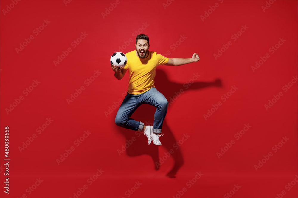Full size body length fun excited overjoyed young bearded man football fan in yellow t-shirt cheer up support favorite team jump hold soccer ball isolated on plain dark red background studio portrait.