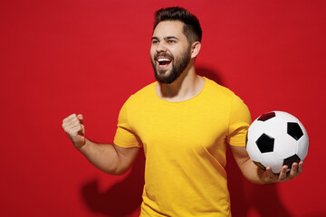 Jubilant fun young bearded man football fan in yellow t-shirt cheer up support favorite team hold soccer ball look aside clenching fists say yes isolated on plain dark red background studio portrait.
