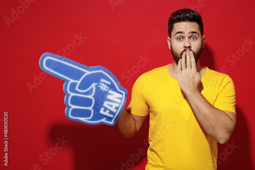 Confused puzzled young bearded man 20s in yellow t-shirt cheer up support favorite team point fan foam glove finger up aside cover mouth with hand isolated plain dark red background studio portrait © ViDi Studio