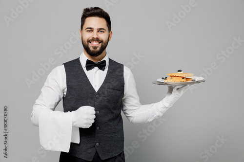 Young happy barista male waiter butler man wear white shirt vest elegant uniform work at cafe hold cheesecake waffles isolated on plain grey background studio portrait. Restaurant employee concept.