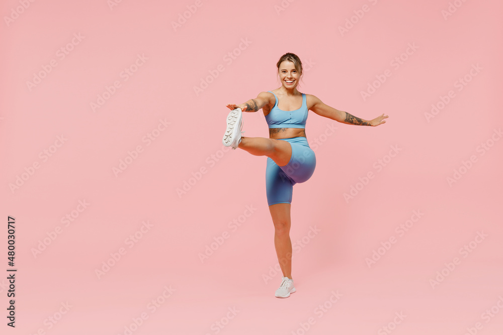 Full body young strong sporty athletic fitness trainer instructor woman wear blue tracksuit spend time in home gym do exercise raise leg isolated on plain light pink background. Workout sport concept