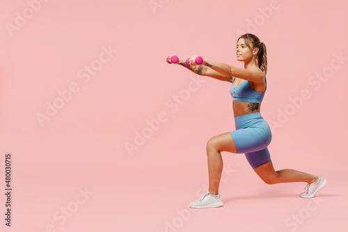 Full body young sporty athletic fitness trainer instructor woman wear blue tracksuit spend time in home gym hold dumbbells do stretch squats isolated on plain pink background. Workout sport concept