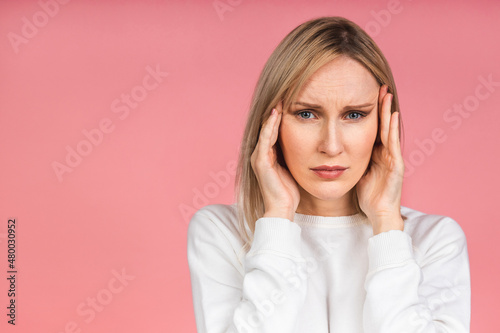 Portrait of a tired middle-aged female blonde woman has a terrible headache, feels pain and frustration, has an unhappy expression, isolated over pink background.
