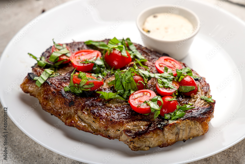 juicy Ribeye steak with fresh tomatoes on white plate on grey concrete table macro close up