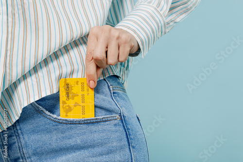 Close up cropped photo shot of caucasian woman wear striped shirt hold in hand credit bank card put into pocket isolated on plain pastel light blue background studio portrait People lifestyle concept photo