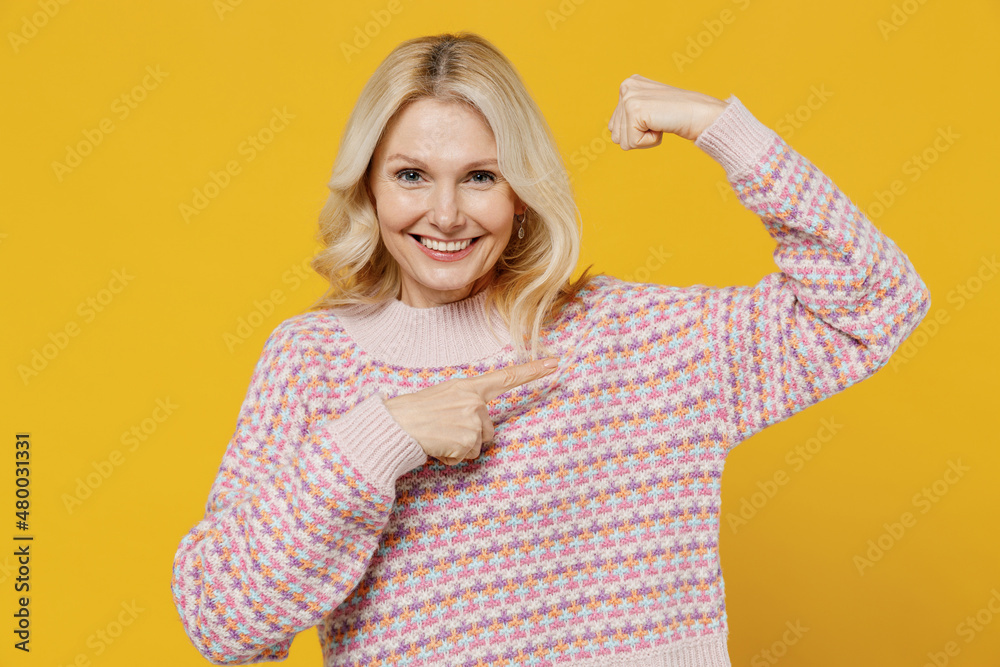 Elderly strong sporty fun fitness woman 50s in pink sweater showing point index finger on biceps muscles on hand demonstrating strength power isolated on plain yellow color background studio portrait. Elderly strong sporty fun fitness woman 50s in pink sweater showing point index finger on biceps muscles on hand demonstrating strength power isolated on plain yellow color background studio portrait.