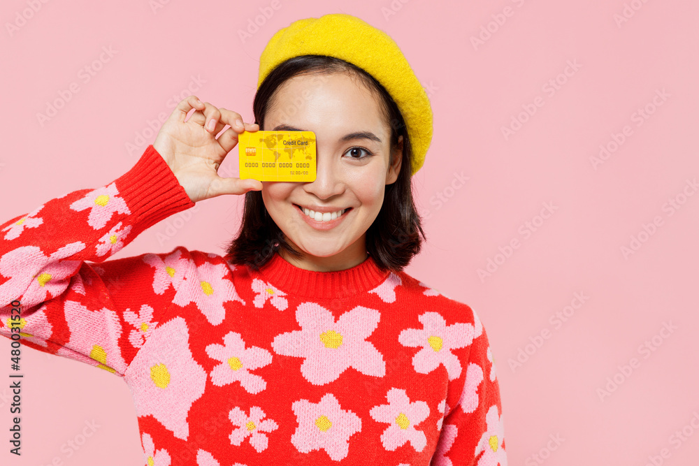 Smiling happy fun woman of Asian ethnicity 20s in red knitted sweater beret cover eye with credit bank card isolated on plain pastel pink background studio portrait. People lifestyle fashion concept Smiling happy fun woman of Asian ethnicity 20s in red knitted sweater beret cover eye with credit bank card isolated on plain pastel pink background studio portrait. People lifestyle fashion concept