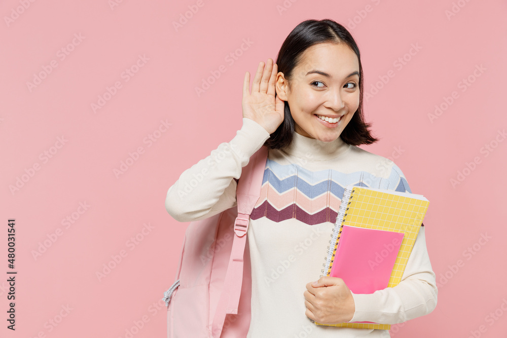 Teen student girl of Asian ethnicity in sweater backpack hold books try to hear you overhear listening intently isolated on pastel plain light pink color background. Education in university concept. Teen student girl of Asian ethnicity in sweater backpack hold books try to hear you overhear listening intently isolated on pastel plain light pink color background. Education in university concept.