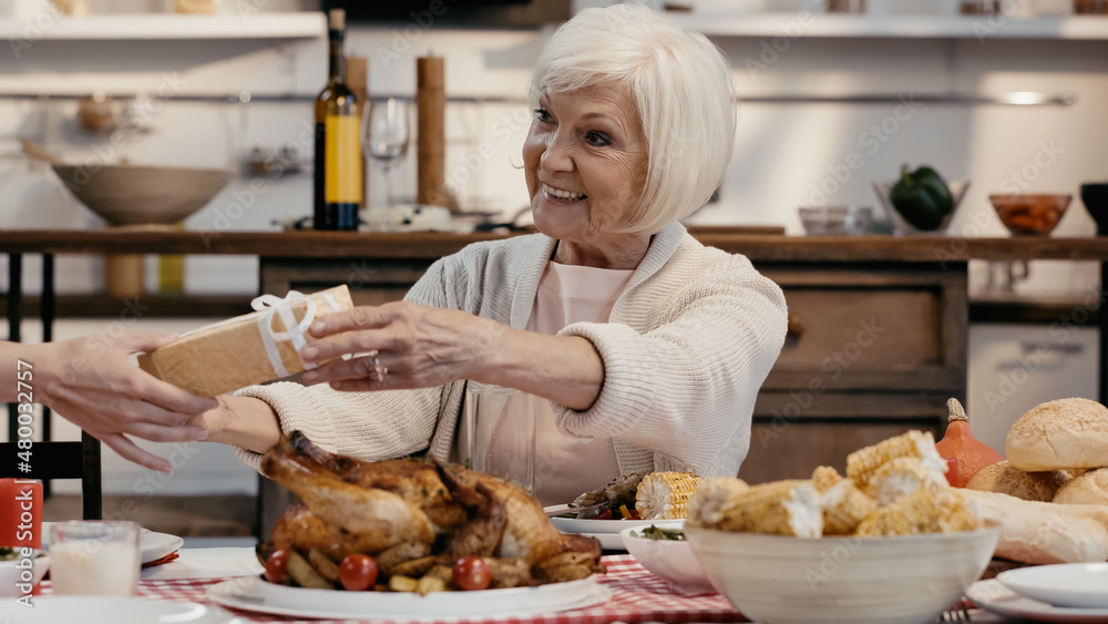 guest giving present to smiling senior woman sitting at table served with thanksgiving dinner.