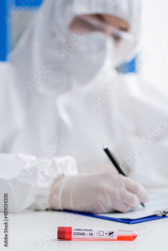 blurred scientist in hazmat suit writing on clipboard near positive covid-19 omicron variant test in lab.