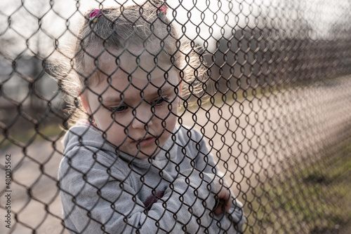 Obraz na plátně A little girl with a sad look behind a metal fence