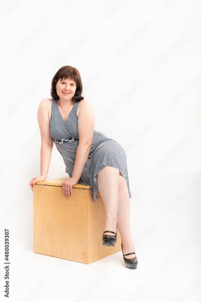 Middle-aged woman with excess weight in dress sitting on a wooden box on white background. Vertical frame Middle-aged woman with excess weight in dress sitting on a wooden box on white background. Vertical frame