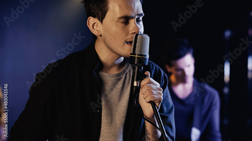 tattooed man singing in microphone near blurred musician on stage.
