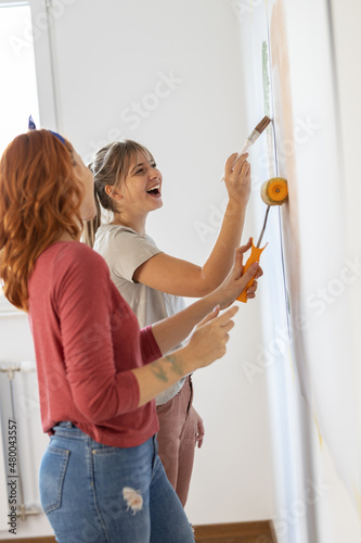 Two female roommates friends using paint roller to decorate walls in they new home.	
