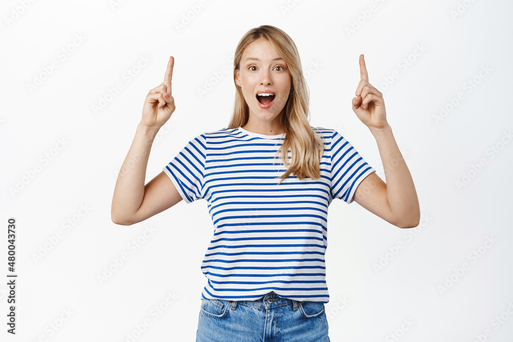 Enthusiastic female model shouts with joy, shows advertisement, points fingers up at top sale promo, stands over white background Enthusiastic female model shouts with joy, shows advertisement, points fingers up at top sale promo, stands over white background