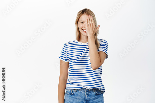 Skin care and women beauty. Happy smiling girl covers half of face, looks with one eye at camera, holds palm on one side of face, standing over white background