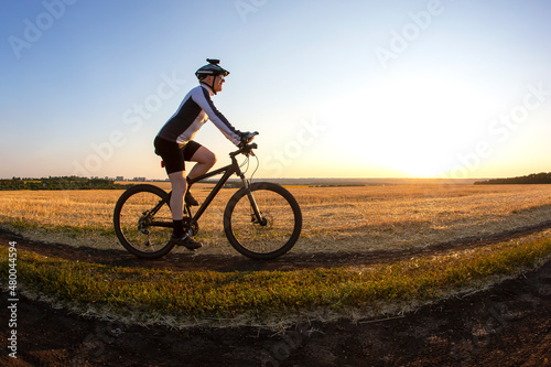 The cyclist rides a bike on the road near the field against the backdrop of the setting sun. Outdoor sports. Healthy lifestyle.