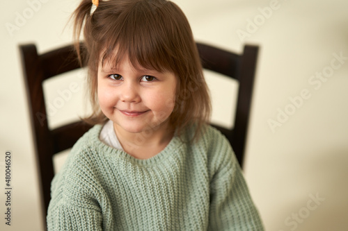 A close-up portrait of a smiling baby with a funny hairstyle and a trick in her eyes. The child is sitting on a chair on a light background.