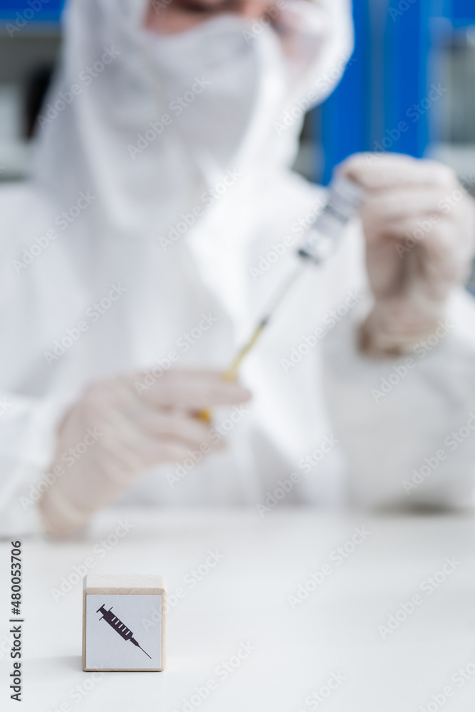 cropped view of blurred immunologist in hazmat suit holding vaccine near cube with syringe icon, omicron variant concept. cropped view of blurred immunologist in hazmat suit holding vaccine near cube with syringe icon, omicron variant concept.