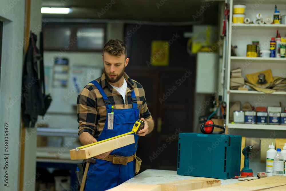 Portrait of young handsome caucasian carpenter who measures a board at the workshop