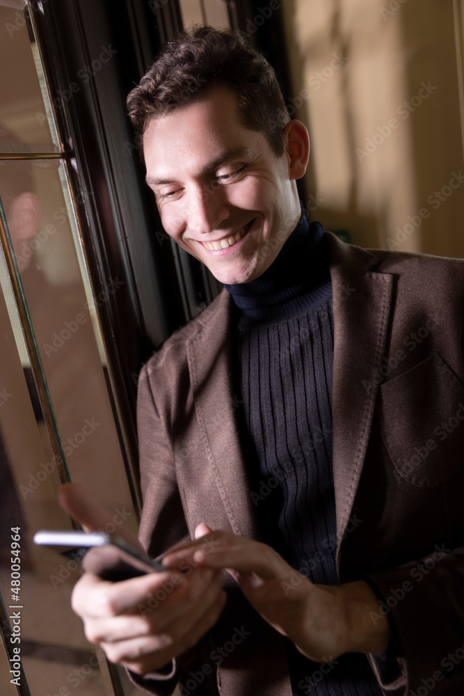 A young man stays indoor texting messages and smiles as he looks at his smartphone A young man stays indoor texting messages and smiles as he looks at his smartphone