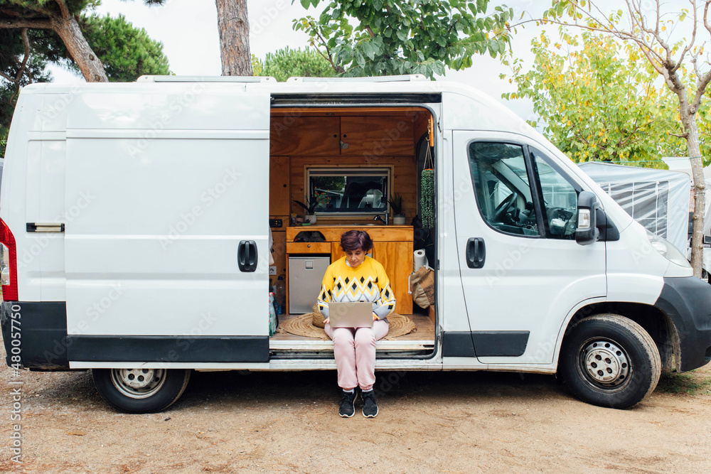 Middle-aged woman sitting with laptop in camper van while working. Nomad life.