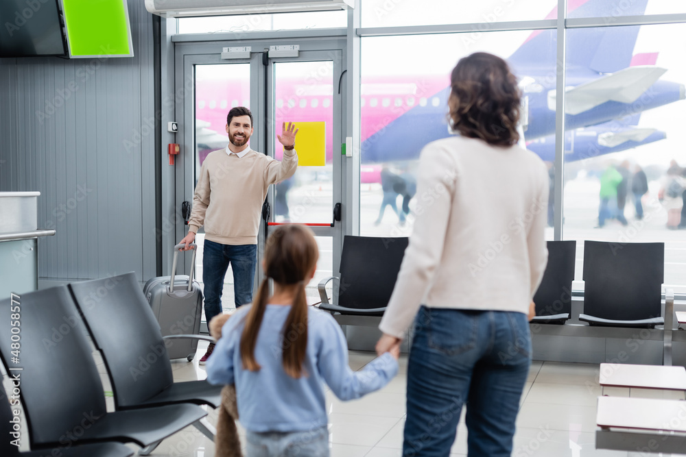 cheerful man with baggage waving hand while looking at wife and daughter in airport. cheerful man with baggage waving hand while looking at wife and daughter in airport.