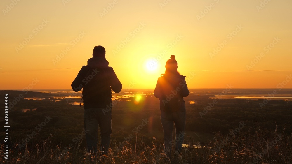 Free travelers, man and woman with backpacks, descend from mountain in rays of dawn, enjoy nature, beautiful sun and landscape. Carefree tourists travel at sunset. Teamwork in business.