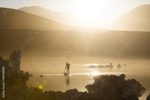 Stand-up paddle-boarder paddles on lake in bright golden sunset flooding scene with golden light. Lake is glistening with light.