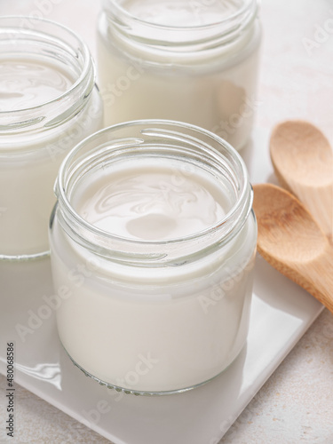Glass jars of homemade plain yogurt with wooden spoons on white tray. Vertical. Close up.