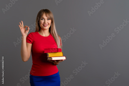 Smart Clever girl holding three books doing ok sign with fingers, smiling friendly gesturing excellent symbol. Young woman with pile color books on grey background. Space for text
