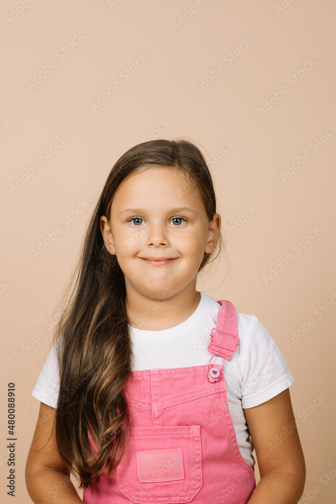Close up face kid with bright shining eyes and calm happy wide smile looking at camera wearing bright, pink jumpsuit and white t-shirt on beige background. Close up face kid with bright shining eyes and calm happy wide smile looking at camera wearing bright, pink jumpsuit and white t-shirt on beige background.