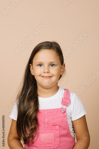 Close up face kid with bright shining eyes and calm happy wide smile looking at camera wearing bright, pink jumpsuit and white t-shirt on beige background.