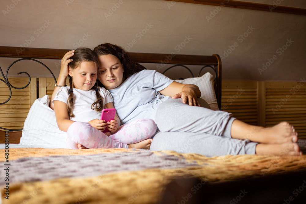 Daughter holding phone in hands with loving mother lying on double bed both looking at phone in wooden house wearing home clothes in daytime. Family time together.
