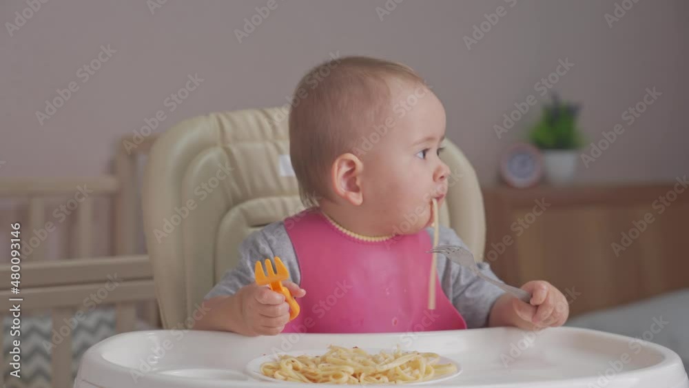 Toddler eats spaghetti for dinner by himself holding two forks in his hands and sitting on a feeding chair Toddler eats spaghetti for dinner by himself holding two forks in his hands and sitting on a feeding chair