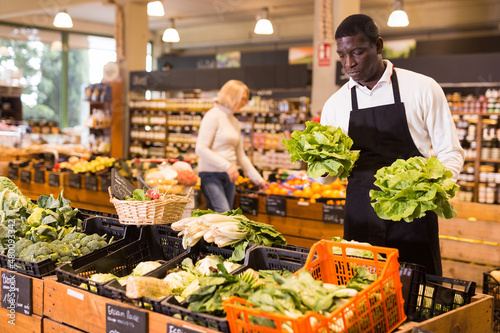 Focused African man working in organic food store, putting fresh vegetables in boxes on showcase