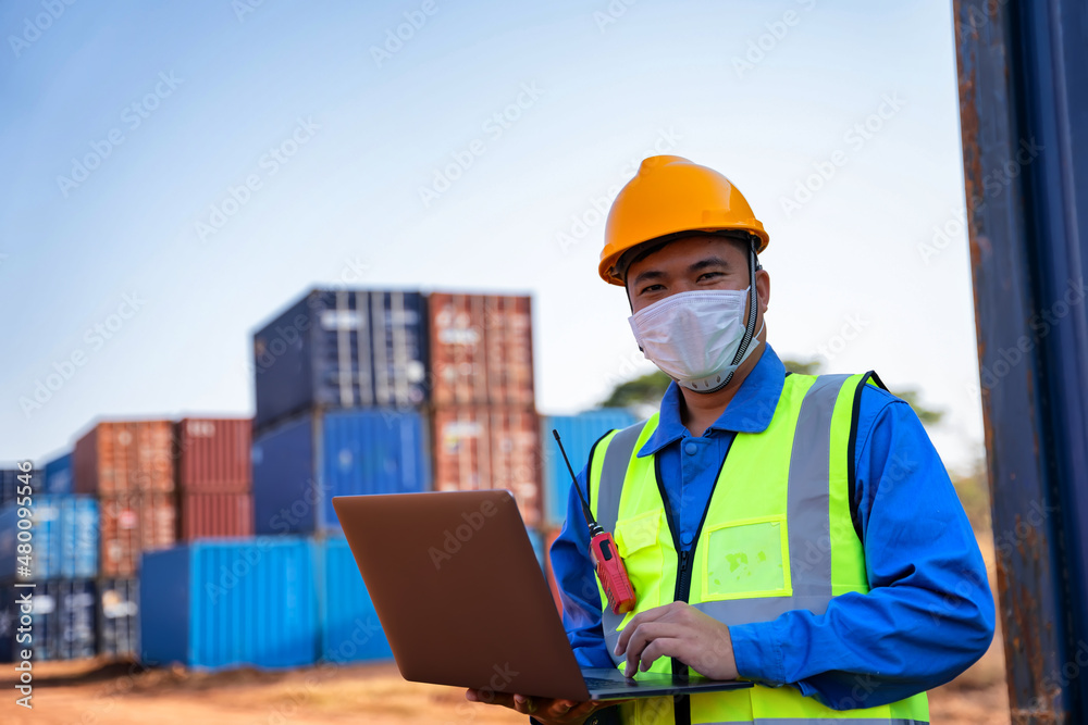 An industrial worker wearing a yellow protective cap holds a laptop to inspect the container yard. An industrial worker wearing a yellow protective cap holds a laptop to inspect the container yard.