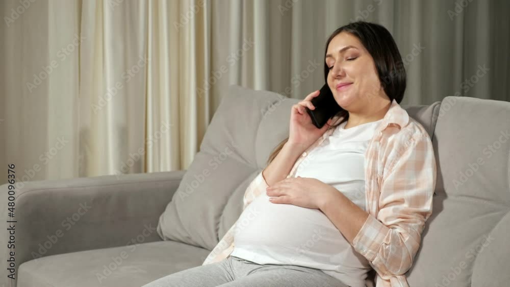Pregnant brunette long-haired woman in flannel shirt talks via cellphone stroking large belly with loving smile sitting on sofa in living room. Pregnant brunette long-haired woman in flannel shirt talks via cellphone stroking large belly with loving smile sitting on sofa in living room.