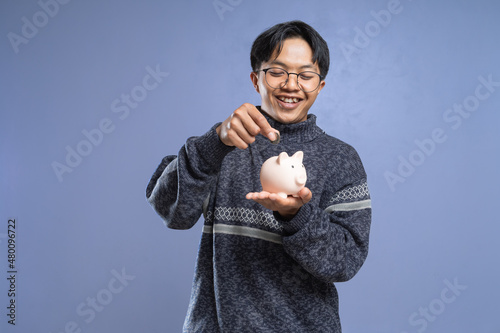 Fototapeta Smiling asian man putting money to his piggy bank