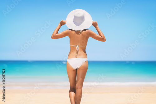 Beach vacation bikini model woman looking at ocean relaxing wearing sun hat. View from behind on summer vacation. Travel wellness skincare healthy body care.